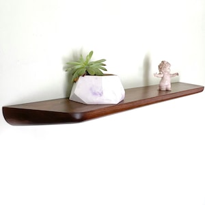 Puede incluir: Una estantería de madera marrón oscuro con bordes redondeados, montada en una pared blanca. Un pequeño macetero geométrico blanco con una planta suculenta verde se encuentra en la estantería, junto con una pequeña figura rosa.