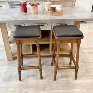 May include: Two wooden bar stools with black leather seats. The stools have a brass footrest and are set against a white kitchen island.