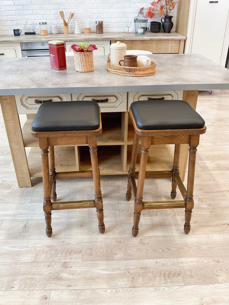 May include: Two wooden bar stools with black leather seats. The stools have a brass footrest and are set against a white kitchen island.