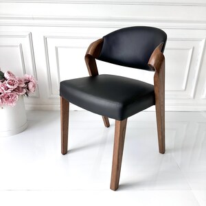 Puede incluir: Silla moderna de cuero negro con respaldo y reposabrazos curvos. La silla tiene estructura y patas de madera, y est&aacute; colocada contra una pared blanca. Un mueble elegante.