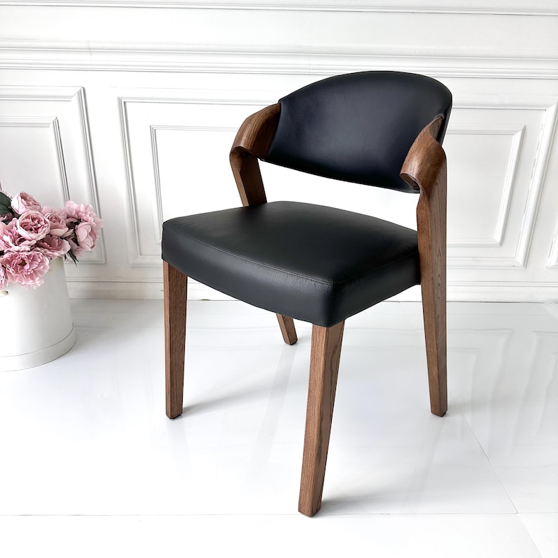 Puede incluir: Silla moderna de cuero negro con respaldo y reposabrazos curvos. La silla tiene estructura y patas de madera, y est&aacute; colocada contra una pared blanca. Un mueble elegante.