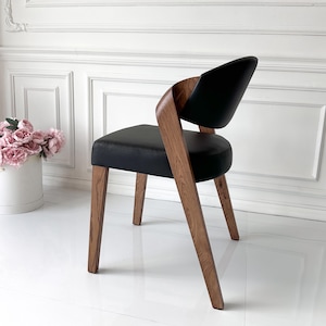 Puede incluir: Silla de comedor moderna con asiento y respaldo de cuero negro. La silla presenta un marco y patas de madera curvada, mostrando un acabado de veta de madera natural. La silla est&aacute; colocada contra una pared y un suelo blancos.