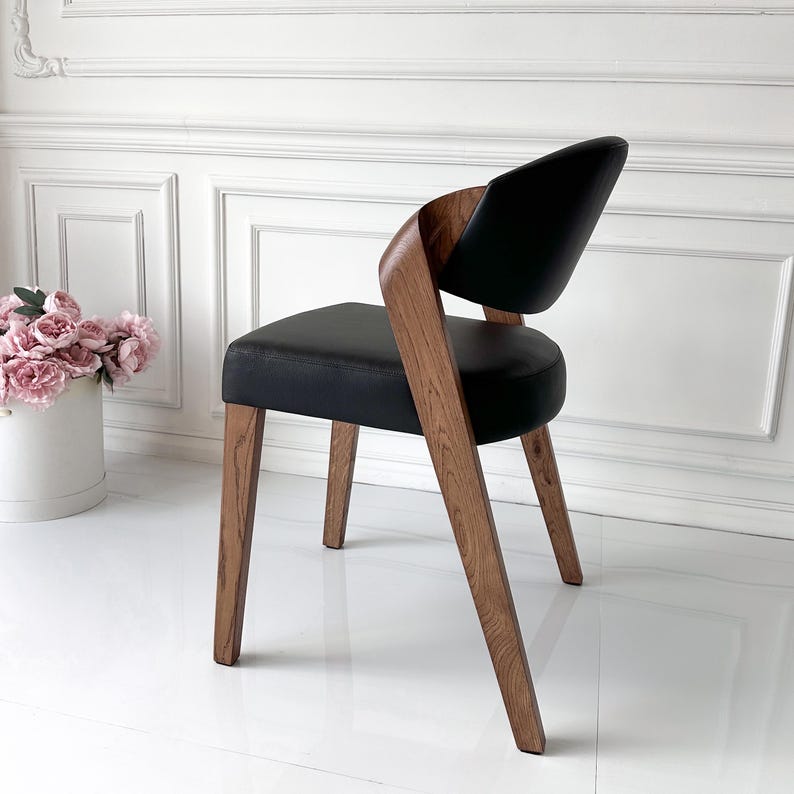 Puede incluir: Silla de comedor moderna con asiento y respaldo de cuero negro. La silla presenta un marco y patas de madera curvada, mostrando un acabado de veta de madera natural. La silla est&aacute; colocada contra una pared y un suelo blancos.