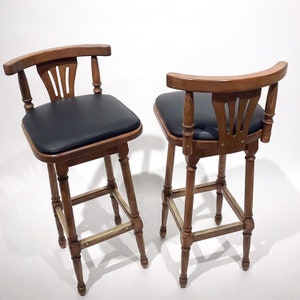 May include: Two wooden bar stools with black leather seats and brass accents. The stools have a curved back and a simple design.