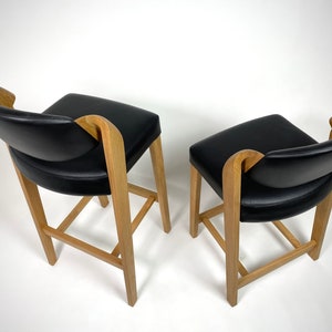 May include: Two black leather bar stools with wooden frames. The stools have a curved back and a simple design.