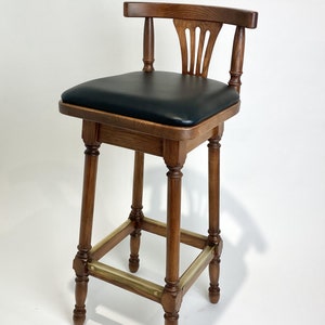 May include: A wooden bar stool with a black leather seat and brass accents. The stool has a traditional design with turned legs and a slatted back.