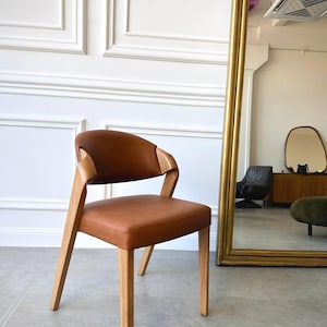 K&ouml;nnte beinhalten: Ein moderner Stuhl mit braunem Ledersitz und R&uuml;ckenlehne. Der Stuhl hat ein helles Holzgestell und Armlehnen. Der Stuhl steht vor einer wei&szlig;en Wand und einem gro&szlig;en goldgerahmten Spiegel.