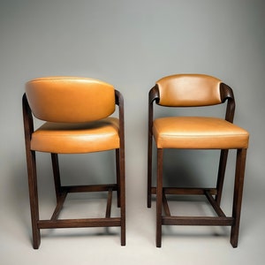 May include: Two brown wooden bar stools with tan leather seats. The stools have a simple, rectangular design with a square base.