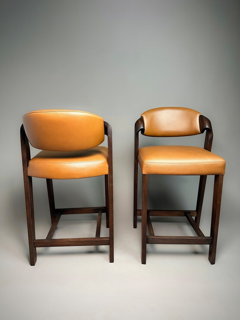 May include: Two brown wooden bar stools with tan leather seats. The stools have a simple, rectangular design with a square base.