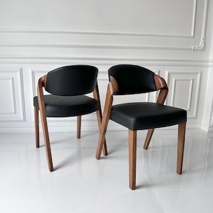 Puede incluir: Dos sillas modernas con asientos y respaldos de cuero negro. Las sillas tienen marcos y patas de madera. Los reposabrazos son de madera y se curvan alrededor del respaldo. Las sillas est&aacute;n colocadas contra una pared blanca.