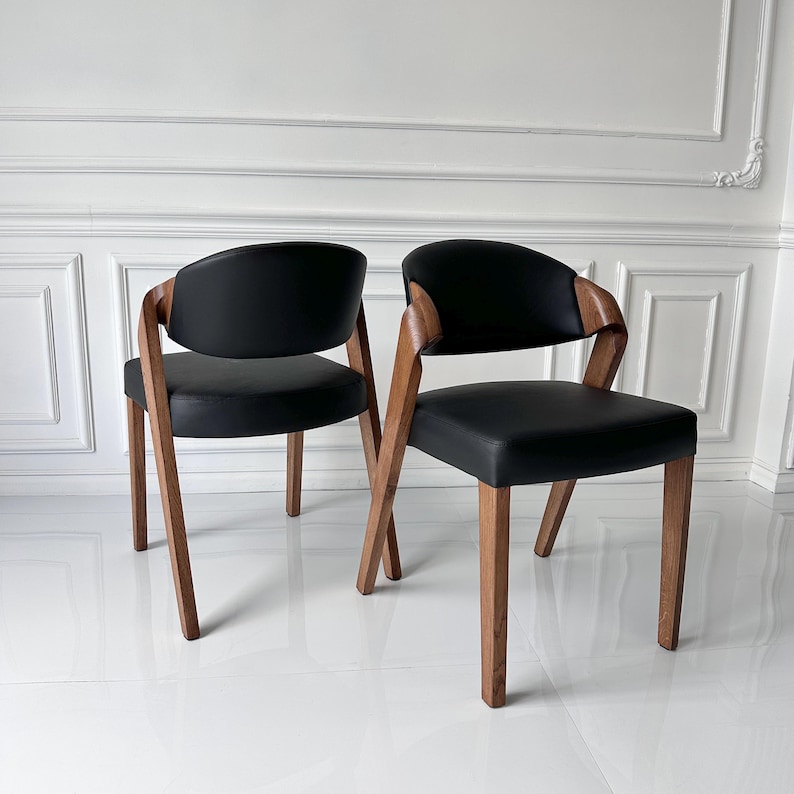 Puede incluir: Dos sillas modernas con asientos y respaldos de cuero negro. Las sillas tienen marcos y patas de madera. Los reposabrazos son de madera y se curvan alrededor del respaldo. Las sillas est&aacute;n colocadas contra una pared blanca.