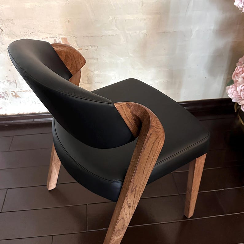 Puede incluir: Silla moderna con asiento y respaldo de cuero negro. Presenta reposabrazos y patas de madera curvados, mostrando un acabado de veta natural. El dise&ntilde;o combina l&iacute;neas elegantes con elementos org&aacute;nicos.