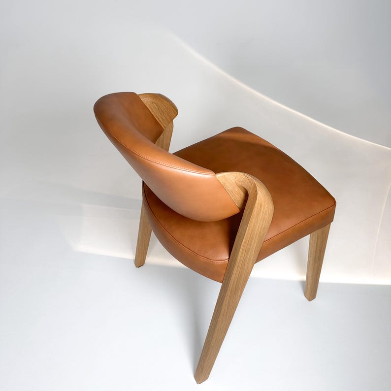 K&ouml;nnte beinhalten: Moderner Stuhl mit geschwungener R&uuml;ckenlehne und Sitzfl&auml;che. Der Stuhl hat eine hellbraune Ledersitzfl&auml;che und R&uuml;ckenlehne sowie Holzbeine und Armlehnen. Das Design ist minimalistisch und elegant, geeignet f&uuml;r Wohn- oder Esszimmer. Ma&szlig;e in Zentimetern.