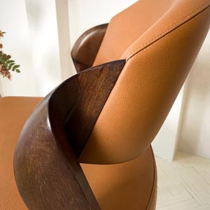 K&ouml;nnte beinhalten: Nahaufnahme eines modernen Stuhls mit geschwungenen Holzarmlehnen und einer Sitzfl&auml;che und R&uuml;ckenlehne aus hellbraunem Leder. Das Holz ist dunkelbraun und das Leder hat sichtbare N&auml;hte.