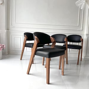 Puede incluir: Cuatro sillas modernas con asientos de cuero negro y reposabrazos y patas de madera curvados. Las sillas tienen un dise&ntilde;o minimalista, con un enfoque en l&iacute;neas limpias y materiales naturales. Las sillas est&aacute;n dispuestas en una habitaci&oacute;n con paredes blancas y suelo blanco brillante.