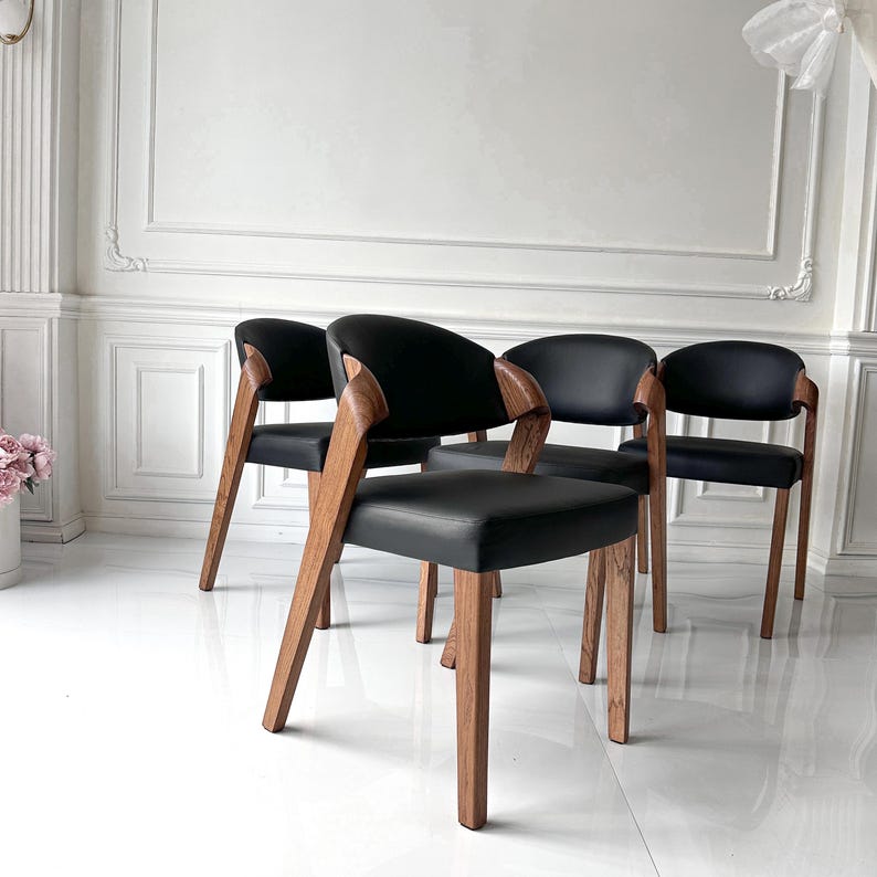 Puede incluir: Cuatro sillas modernas con asientos de cuero negro y reposabrazos y patas de madera curvados. Las sillas tienen un dise&ntilde;o minimalista, con un enfoque en l&iacute;neas limpias y materiales naturales. Las sillas est&aacute;n dispuestas en una habitaci&oacute;n con paredes blancas y suelo blanco brillante.