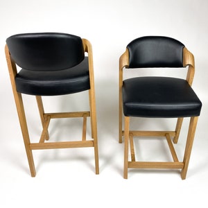 May include: Two black leather bar stools with wooden frames. The stools have a simple design with a rectangular seat and a low back.
