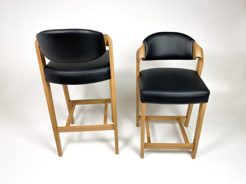 May include: Two black leather bar stools with wooden frames. The stools have a simple design with a rectangular seat and a low back.