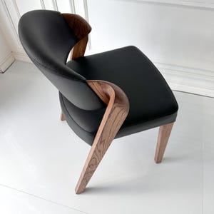 Puede incluir: Silla moderna de cuero negro con reposabrazos y patas de madera curvados. La silla tiene un asiento cuadrado y un respaldo curvo. La madera presenta un veteado natural. La silla est&aacute; dise&ntilde;ada para la comodidad y el estilo.