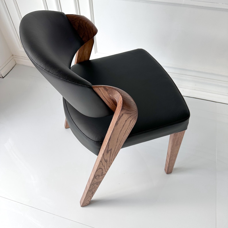 Puede incluir: Silla moderna de cuero negro con reposabrazos y patas de madera curvados. La silla tiene un asiento cuadrado y un respaldo curvo. La madera presenta un veteado natural. La silla est&aacute; dise&ntilde;ada para la comodidad y el estilo.