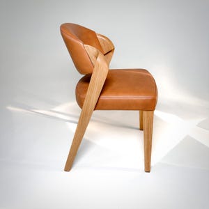 K&ouml;nnte beinhalten: Moderner Stuhl mit hellbraunem Ledersitz und R&uuml;ckenlehne. Der Stuhl hat hellfarbene Holzbeine und eine einzigartige, geschwungene Holzr&uuml;ckenlehne. Das Design ist minimalistisch und elegant, geeignet f&uuml;r den Heim- oder B&uuml;rogebrauch. Ma&szlig;e in Zentimetern.