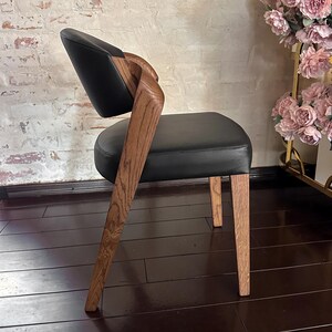 Puede incluir: Silla de comedor moderna con asiento y respaldo de cuero negro. La silla presenta un reposabrazos y patas de madera curvados, mostrando un acabado de veta natural. El dise&ntilde;o de la silla es elegante y contempor&aacute;neo.