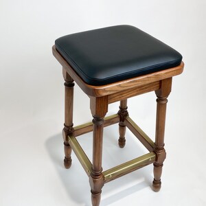 May include: A wooden bar stool with a black leather seat and brass accents. The stool has a square base and turned legs.