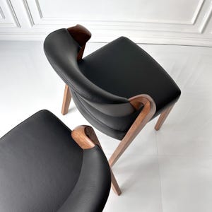 Puede incluir: Dos sillones modernos con asientos de cuero negro y reposabrazos curvos de madera. Las sillas tienen un dise&ntilde;o minimalista con patas de madera en &aacute;ngulo, sobre un fondo blanco. Las sillas son de color marr&oacute;n oscuro.