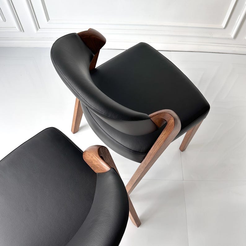 Puede incluir: Dos sillones modernos con asientos de cuero negro y reposabrazos curvos de madera. Las sillas tienen un dise&ntilde;o minimalista con patas de madera en &aacute;ngulo, sobre un fondo blanco. Las sillas son de color marr&oacute;n oscuro.