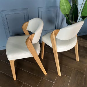 Könnte beinhalten: Zwei moderne Sessel mit hellgrauen Ledersitzen und geschwungenen Armlehnen und Beinen aus Holz. Die Stühle haben ein minimalistisches Design mit Fokus auf natürlichen Materialien und klaren Linien. Der Hintergrund ist eine blaue Wand und dunkler Holzboden.