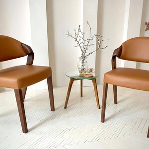 K&ouml;nnte beinhalten: Zwei moderne Sessel mit braunen Ledersitzen und dunklen Holzrahmen. Die St&uuml;hle haben geschwungene R&uuml;ckenlehnen und Armlehnen. Zwischen den St&uuml;hlen steht ein kleiner Beistelltisch mit Glasplatte und Holzbeinen. Auf dem Tisch steht eine Vase mit Zweigen.