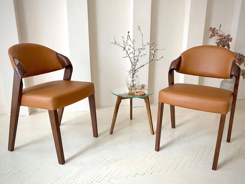 K&ouml;nnte beinhalten: Zwei moderne Sessel mit braunen Ledersitzen und dunklen Holzrahmen. Die St&uuml;hle haben geschwungene R&uuml;ckenlehnen und Armlehnen. Zwischen den St&uuml;hlen steht ein kleiner Beistelltisch mit Glasplatte und Holzbeinen. Auf dem Tisch steht eine Vase mit Zweigen.