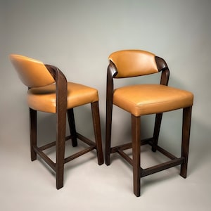 May include: Two brown wooden bar stools with tan leather seats. The stools have a simple design with a curved back and a footrest.