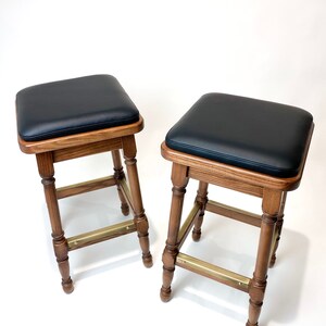 May include: Two wooden bar stools with black leather seats and brass accents. The stools have a traditional design with turned legs and a square seat.