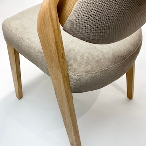 May include: A light brown wooden chair with a beige fabric seat and back. The chair has a unique curved back design and four legs.