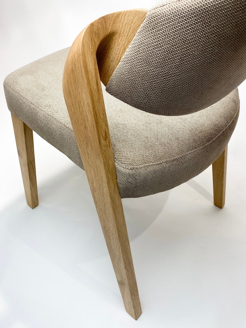 May include: A light brown wooden chair with a beige fabric seat and back. The chair has a unique curved back design and four legs.