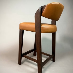 May include: A brown wooden bar stool with a tan leather seat and back. The stool has a simple, modern design with a square base and a curved back.