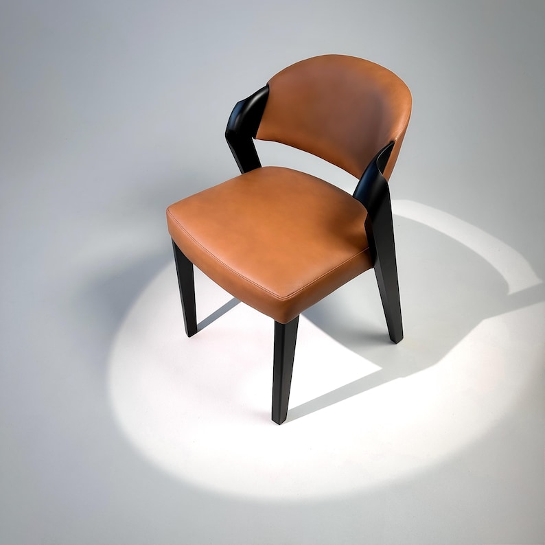 K&ouml;nnte beinhalten: Moderner Stuhl mit Sitzfl&auml;che und R&uuml;ckenlehne aus hellbraunem Leder. Er hat schwarze Holzbeine und Armlehnen. Das Design zeichnet sich durch eine geschwungene R&uuml;ckenlehne und eine quadratische Sitzfl&auml;che aus. Der Stuhl steht vor wei&szlig;em Hintergrund.