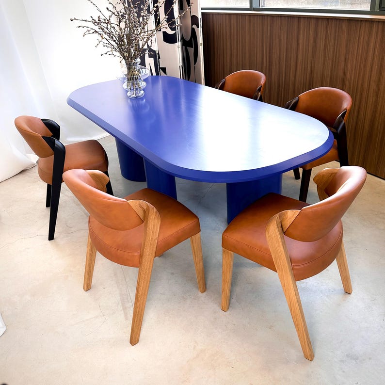 K&ouml;nnte beinhalten: Ein modernes Esszimmer-Set mit einem ovalen blauen Tisch und sechs braunen Lederst&uuml;hlen. Die St&uuml;hle haben Holzbeine und Armlehnen. Eine Vase mit Zweigen steht auf dem Tisch und schafft einen stilvollen Essbereich.