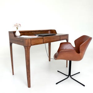 May include: A wooden desk with a laptop, a small white vase with flowers, and a brown leather chair. The desk has a dark wood finish and a curved design. The chair has a black base.