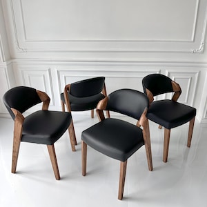 Puede incluir: Cuatro sillas de comedor modernas con asientos de cuero negro y reposabrazos y patas de madera curvados. Las sillas tienen un dise&ntilde;o minimalista, con un enfoque en l&iacute;neas limpias y materiales naturales. Las sillas est&aacute;n dispuestas en grupo.