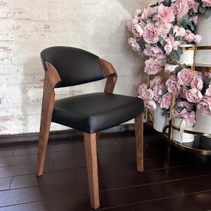 Puede incluir: Una silla negra moderna con respaldo y asiento curvos. La silla tiene patas y reposabrazos de madera. La silla est&aacute; colocada contra una pared de ladrillo y una exhibici&oacute;n de flores rosas.