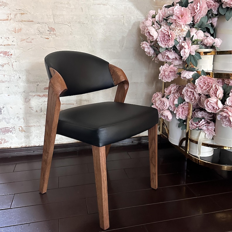 Puede incluir: Una silla negra moderna con respaldo y asiento curvos. La silla tiene patas y reposabrazos de madera. La silla est&aacute; colocada contra una pared de ladrillo y una exhibici&oacute;n de flores rosas.