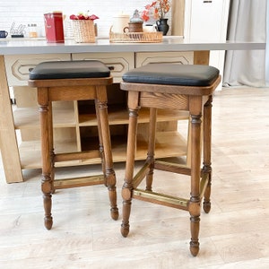 May include: Two wooden bar stools with black leather seats and brass accents. The stools have a simple, classic design and are perfect for a kitchen or bar.