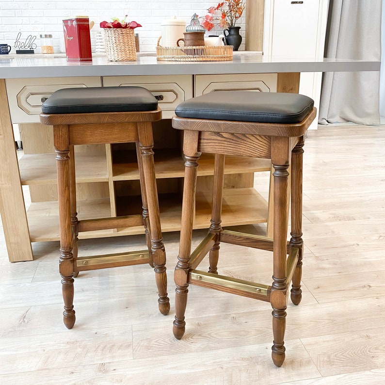 May include: Two wooden bar stools with black leather seats and brass accents. The stools have a simple, classic design and are perfect for a kitchen or bar.