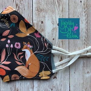 Pode incluir: Uma máscara facial preta com um padrão floral que apresenta uma raposa, pássaros e folhas. A máscara tem tiras ajustáveis brancas. A máscara é feita por Tigerlily By Helen.