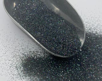 May include: A scoop filled with black glitter with iridescent green and blue flecks. The glitter is spilling out of the scoop onto a white surface.