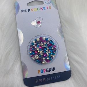 May include: A PopGrip phone grip with a clear background and a colorful, sparkly design. The grip is expandable and has a swappable top. The packaging features the PopSockets logo and the words "PopGrip Premium".