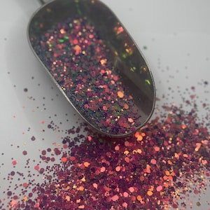Peut inclure: Une cuillère en argent remplie de paillettes irisées rose, vertes et dorées. Les paillettes sont éparpillées sur une surface blanche.