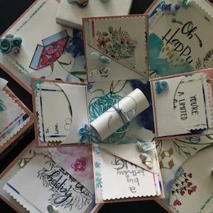 May include: A pink and white explosion box with a watercolour floral design. The box has six sides, each with a different design. The box is decorated with paper flowers, ribbons, and a rolled up message. The text on the box says "Oh Happy", "You're A Limited", "To", "Love", "Happy Birthday", and "You're So Special".
